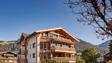 Modernes Chalet-Gebäude mit Holzfassade und Balkonen vor alpiner Bergkulisse unter blauem Himmel.