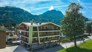 Modernes Apartmentgebäude mit Holzfassade und Balkonen, eingebettet in eine grüne Berglandschaft.