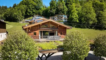 Charmantes Holzchalet in alpiner Umgebung mit grünen Hügeln und Bäumen unter blauem Himmel.