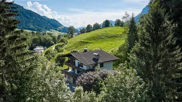 Idyllisches Haus in den Bergen, umgeben von grünen Hügeln und Bäumen unter blauem Himmel.