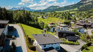 Luftaufnahme eines modernen Hauses in einer malerischen Berglandschaft mit umliegenden Gebäuden und viel Grün.