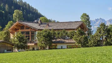 Großes Chalet im alpinen Stil mit Holzfassade und Satteldach, umgeben von grünen Wiesen und Bergen.