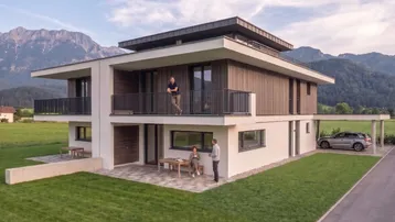 Modernes Einfamilienhaus mit Balkonen, Terrasse und Carport vor alpiner Bergkulisse.