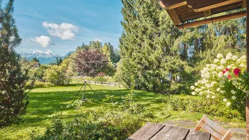 Gemütlicher Außenbereich mit Holztisch und Stühlen, umgeben von einem grünen Garten mit Bergblick.