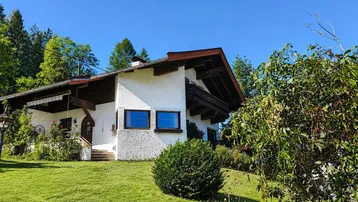 Weißes Haus im alpinen Stil mit Satteldach und gepflegtem Vorgarten unter blauem Himmel.