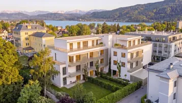 Moderne Wohnanlage mit weißer Fassade, umgeben von Grünflächen und mit Blick auf den See und die Berge.