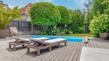 Großer Gartenbereich mit Holzterrasse, Swimmingpool und Liegestühlen unter blauem Himmel.