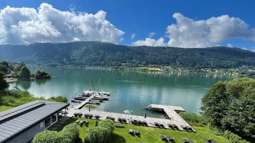 Panoramablick auf den See mit Bootsstegen, Liegewiese und umliegenden Bergen unter blauem Himmel.