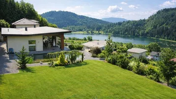 Panoramablick auf das moderne Haus mit großem Garten, Naturpool und See im Hintergrund.
