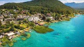 Panoramablick auf eine malerische Siedlung am Seeufer mit bewaldeten Bergen im Hintergrund.