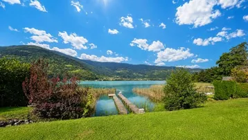 Idyllischer Blick auf den See mit Bergen, einem Holzsteg und grüner Wiese unter strahlendem Himmel.