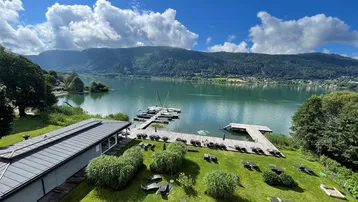 Panoramablick auf den See mit Bootsstegen, Liegewiese und umliegenden Bergen unter blauem Himmel.