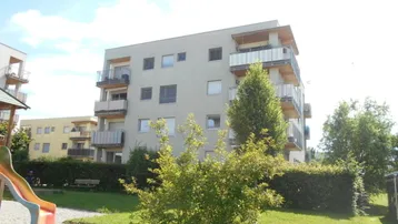 Modernes Mehrfamilienhaus mit Balkonen und einem Spielplatz im Vordergrund unter blauem Himmel.