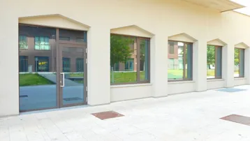 Eingangsbereich eines modernen Gebäudes mit großen Glasfenstern und einer gepflasterten Fläche davor.
