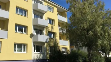 Gelbe Fassade eines Mehrfamilienhauses mit Balkonen, großen Fenstern und einem Baum im Vordergrund.