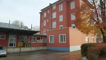 Rotes Mehrfamilienhaus mit mehreren Stockwerken, Parkplatz und herbstlicher Baumkulisse.