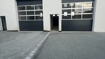 Moderne Gebäudefassade mit zwei großen Garagentoren und separatem Eingangsbereich, davor eine asphaltierte Zufahrt.