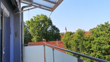 Balkon mit Blick auf grüne Bäume, Dächer und einen Kirchturm in der Ferne.