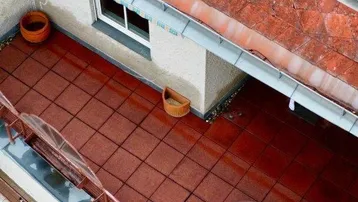 Dachgeschoss-Terrasse mit roten Fliesen und einem kleinen Tisch, umgeben von Ziegeldächern in der Stadt.