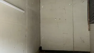Leerer Raum mit Betonboden und einfachen Wandpaneelen, beleuchtet durch Neonröhren an Decke und Wand.