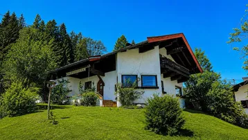 Traditionelles Haus auf einem grünen Hügel, umgeben von Bäumen unter strahlend blauem Himmel.