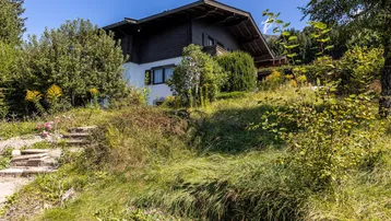 Charmantes Haus im alpinen Stil auf einem Hanggrundstück mit üppiger Vegetation und Steintreppe.