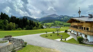 Traditionelles Chalet-Gebäude in alpiner Landschaft mit weiten grünen Wiesen und Bergblick unter bewölktem Himmel.