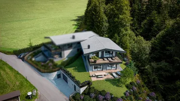 Luftaufnahme eines modernen Hauses in alpiner Landschaft mit Terrassen und viel Grün.