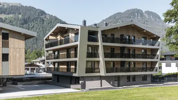 Modernes Apartmentgebäude mit Holzfassade und Balkonen vor alpiner Bergkulisse.