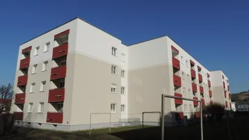 Modernes Mehrfamilienhaus mit weißer und beiger Fassade sowie roten Balkonen unter blauem Himmel.