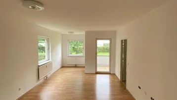 Geräumiger, leerer Wohnbereich mit hellem Laminatboden und Zugang zum Balkon.
