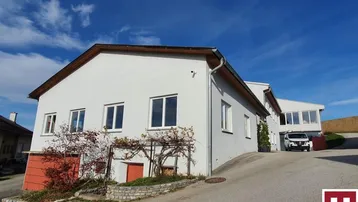 Ansicht des weißen Gebäudes mit Zufahrt und grüner Umgebung unter blauem Himmel.