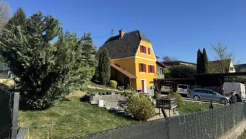 Gelbes Haus mit rotem Dach und Fensterläden, umgeben von Bäumen und einem Gartenbereich.