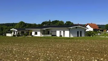 Modernes Einfamilienhaus mit weißer Fassade und dunklem Dach, umgeben von Feldern und Natur.