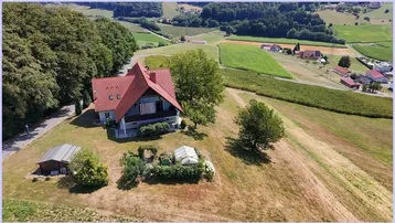 Luftaufnahme eines Hauses mit rotem Dach, umgeben von grünen Hügeln und Feldern in ländlicher Umgebung.