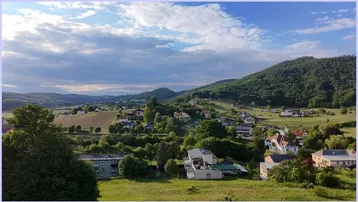 Weitläufige Landschaftsaufnahme eines Dorfes in einem grünen Tal mit bewaldeten Hügeln unter bewölktem Himmel.