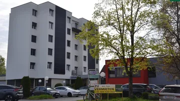 Mehrstöckiges Gebäude mit weißer und schwarzer Fassade, umgeben von einem Parkplatz und Bäumen.