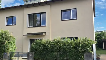 Gepflegtes Einfamilienhaus mit beigefarbener Fassade, großen Fenstern und grüner Hecke im Vorgarten.