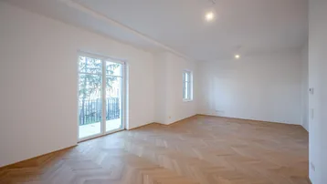 Helles, unmöbliertes Wohnzimmer mit Fischgrätparkett und Zugang zu einem Balkon.