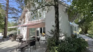 Große Terrasse mit Gartenmöbeln vor der modernen Villa, umgeben von Bäumen und Natur.