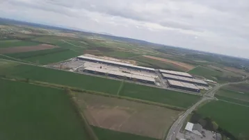 Luftaufnahme von großen Industriegebäuden umgeben von weiten Feldern und Straßen unter bewölktem Himmel.