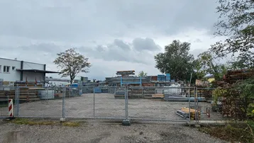 Außenbereich mit Bauzäunen und gelagerten Materialien unter bewölktem Himmel, angrenzend an ein Gebäude.