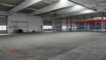 Große, leere Lagerhalle mit Betonboden, hohen Decken und Tageslicht durch Oberlichter und Rolltore.