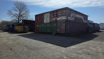 Industrielles Außengelände mit gestapelten Schiffscontainern und einer großen asphaltierten Fläche unter blauem Himmel.