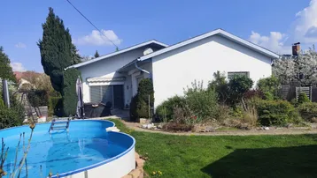 Weißes Einfamilienhaus mit großem Garten und einem oberirdischen Pool unter blauem Himmel.