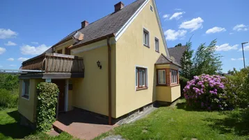 Gelbes Einfamilienhaus mit Balkon und blühendem Rhododendron im Vorgarten.