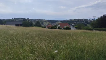 Weitläufiges Feld mit Blick auf eine ländliche Siedlung und hügelige Landschaft unter bewölktem Himmel.