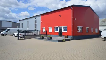 Modernes Gewerbegebäude mit roter und grauer Fassade sowie gepflastertem Vorplatz unter blauem Himmel.
