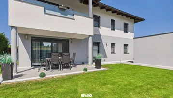 Moderne Hausfassade mit gepflasterter Terrasse, Gartenmöbeln und gepflegtem Rasenbereich.