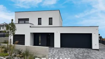 Moderne weiße Villa mit schwarzem Garagentor und gepflasterter Auffahrt unter blauem Himmel.